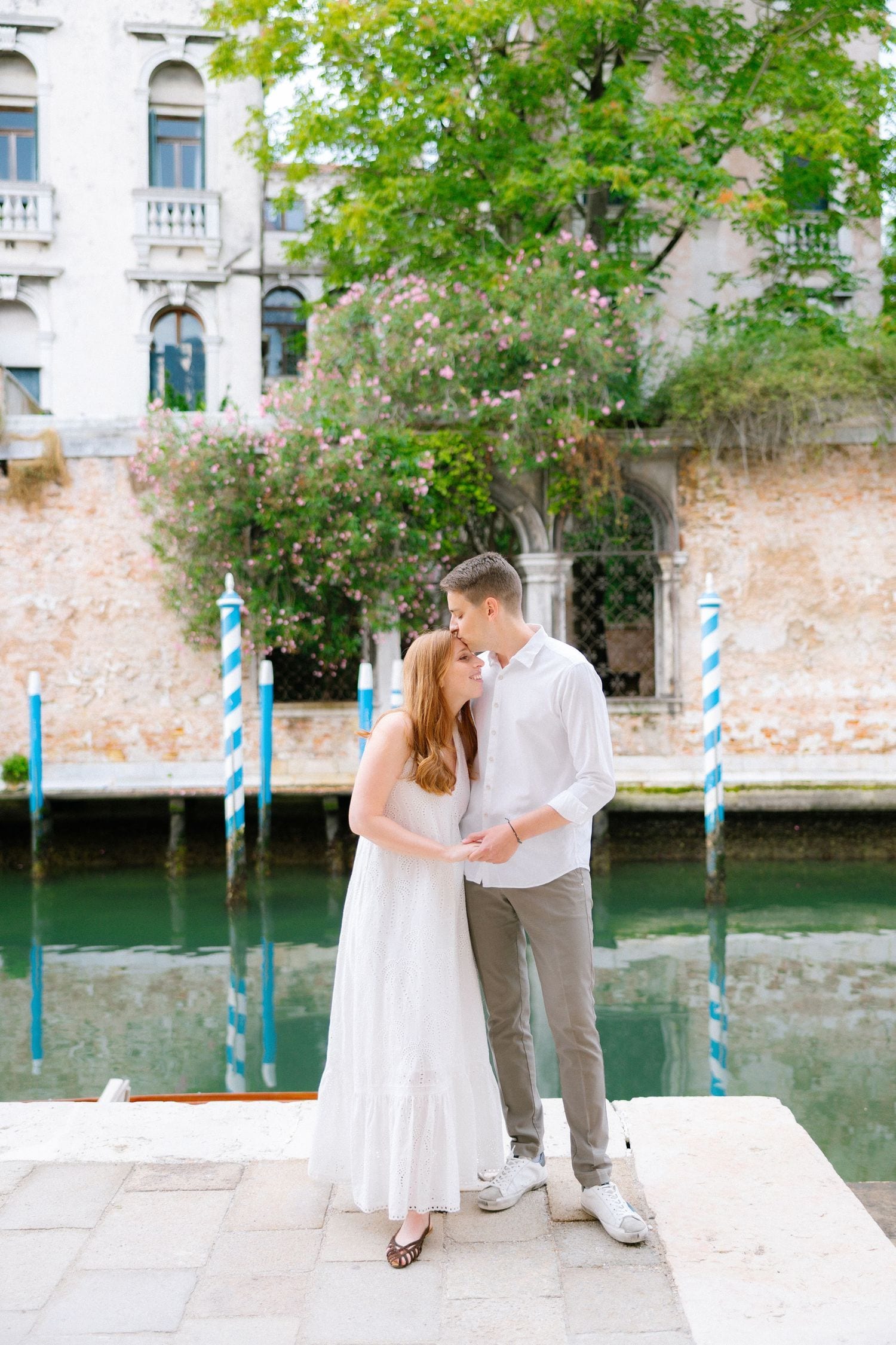 A couple stands together on a dock with blue mooring poles and historic buildings in Venice.