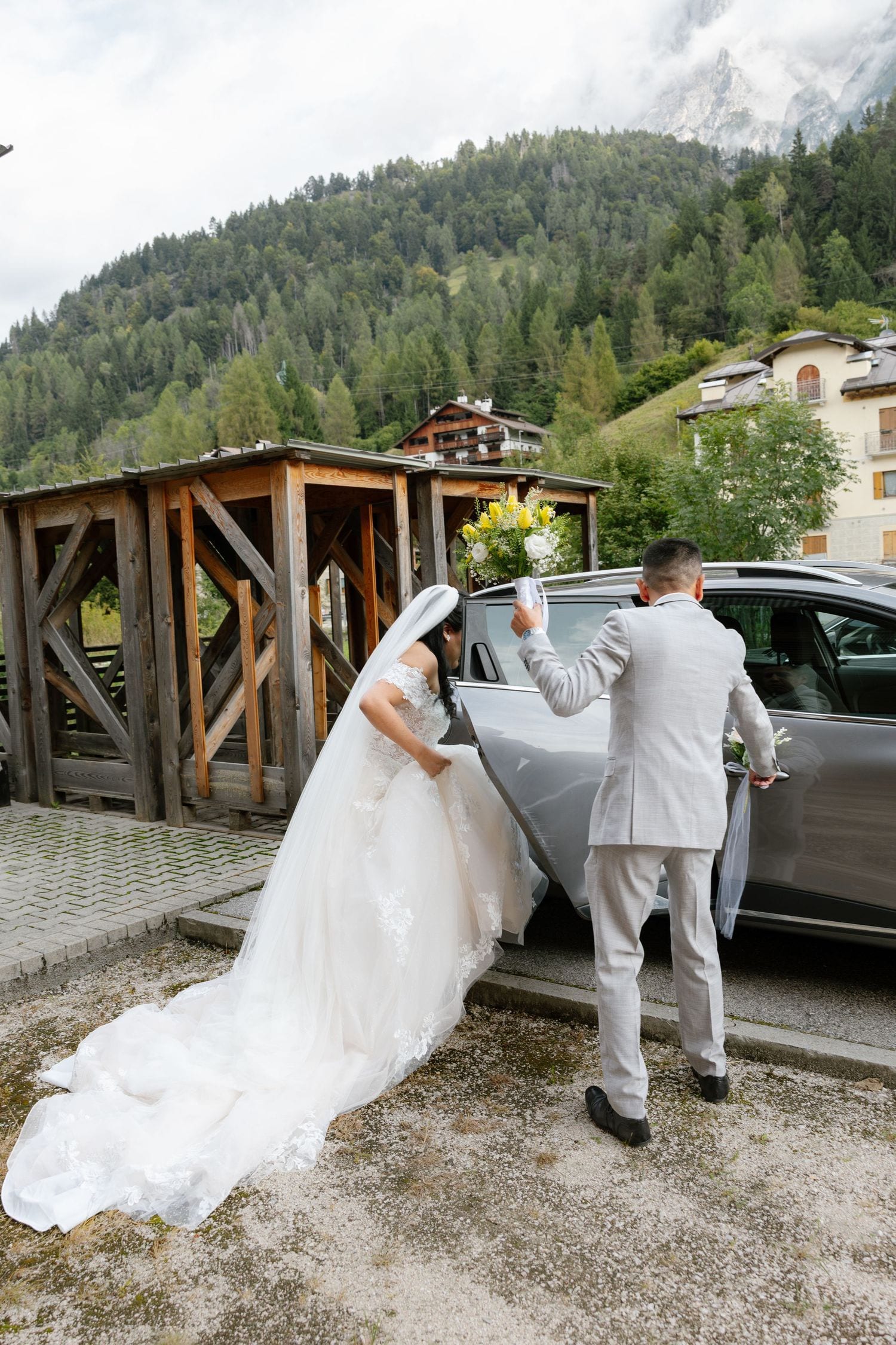 Bride in white gown and groom in gray suit arrive at mountain wedding venue beside luxury car.