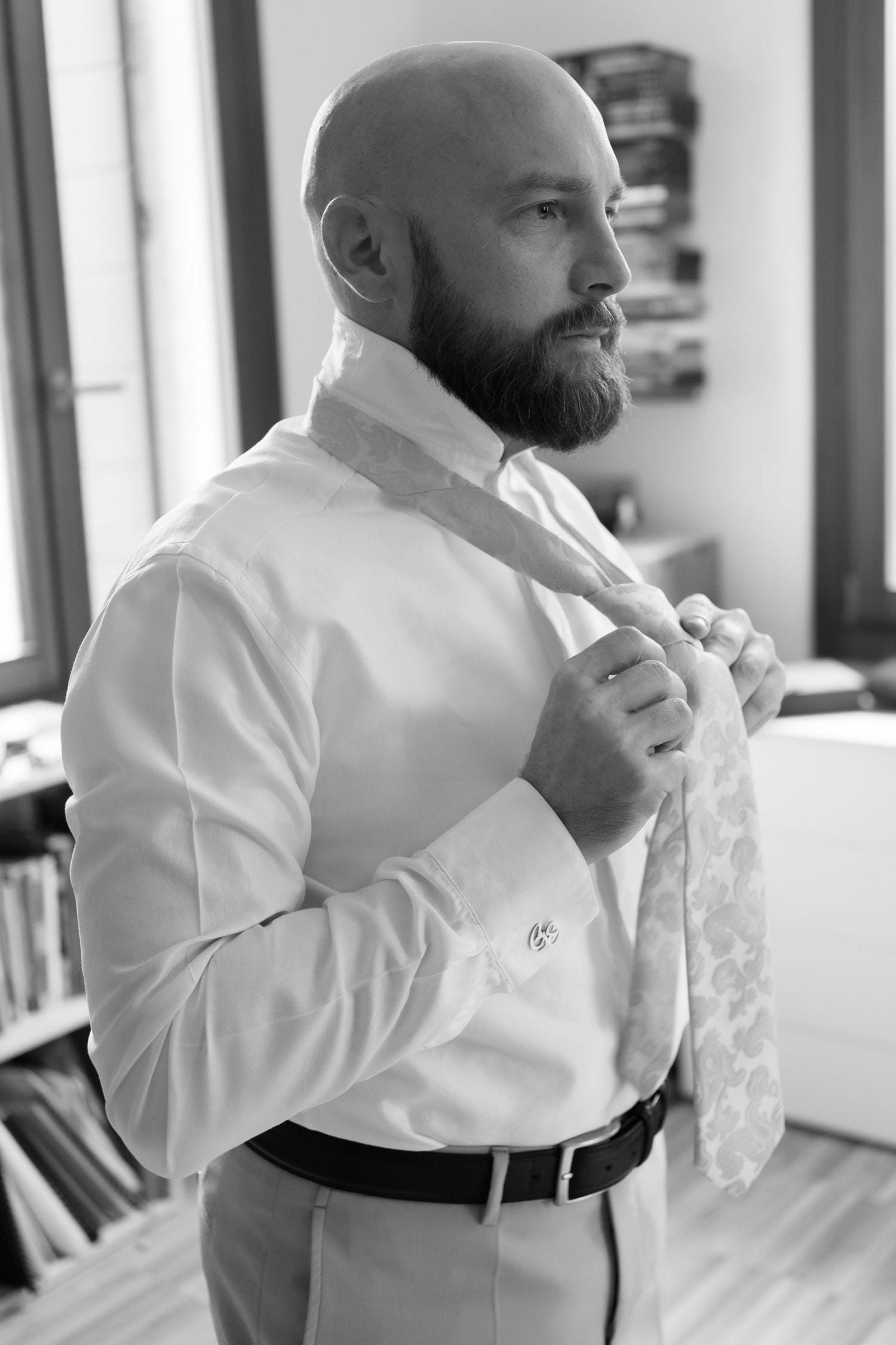 A bearded man in a white dress shirt adjusts his tie while getting ready for a formal occasion.