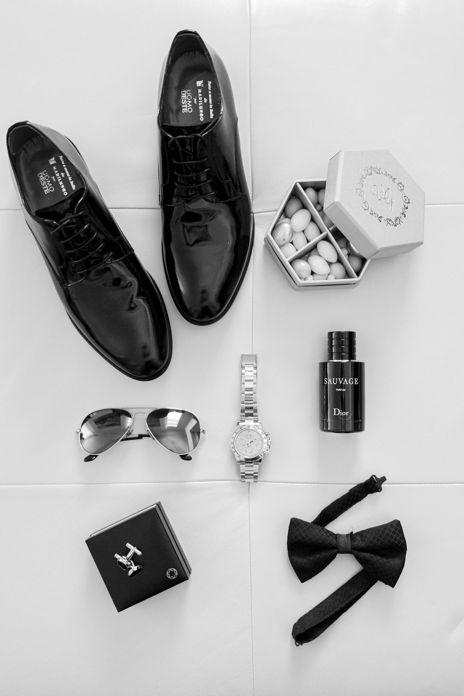 Flat lay of men's wedding accessories including black patent shoes, sunglasses, watch, cologne, bow tie and cuff links.