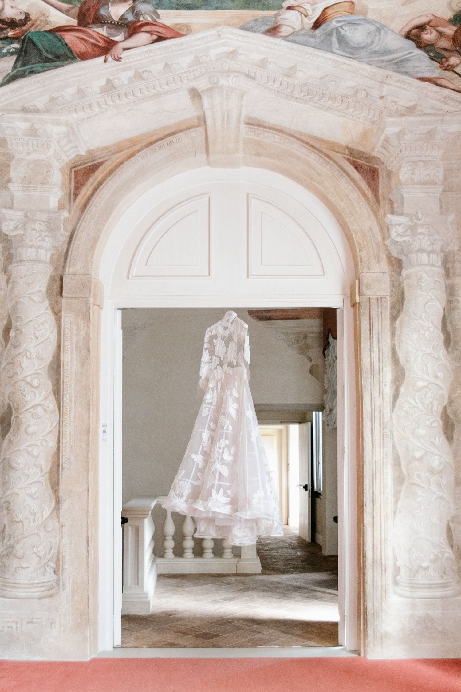 A white lace wedding dress hangs in an ornate archway with stone columns and classical architectural details.