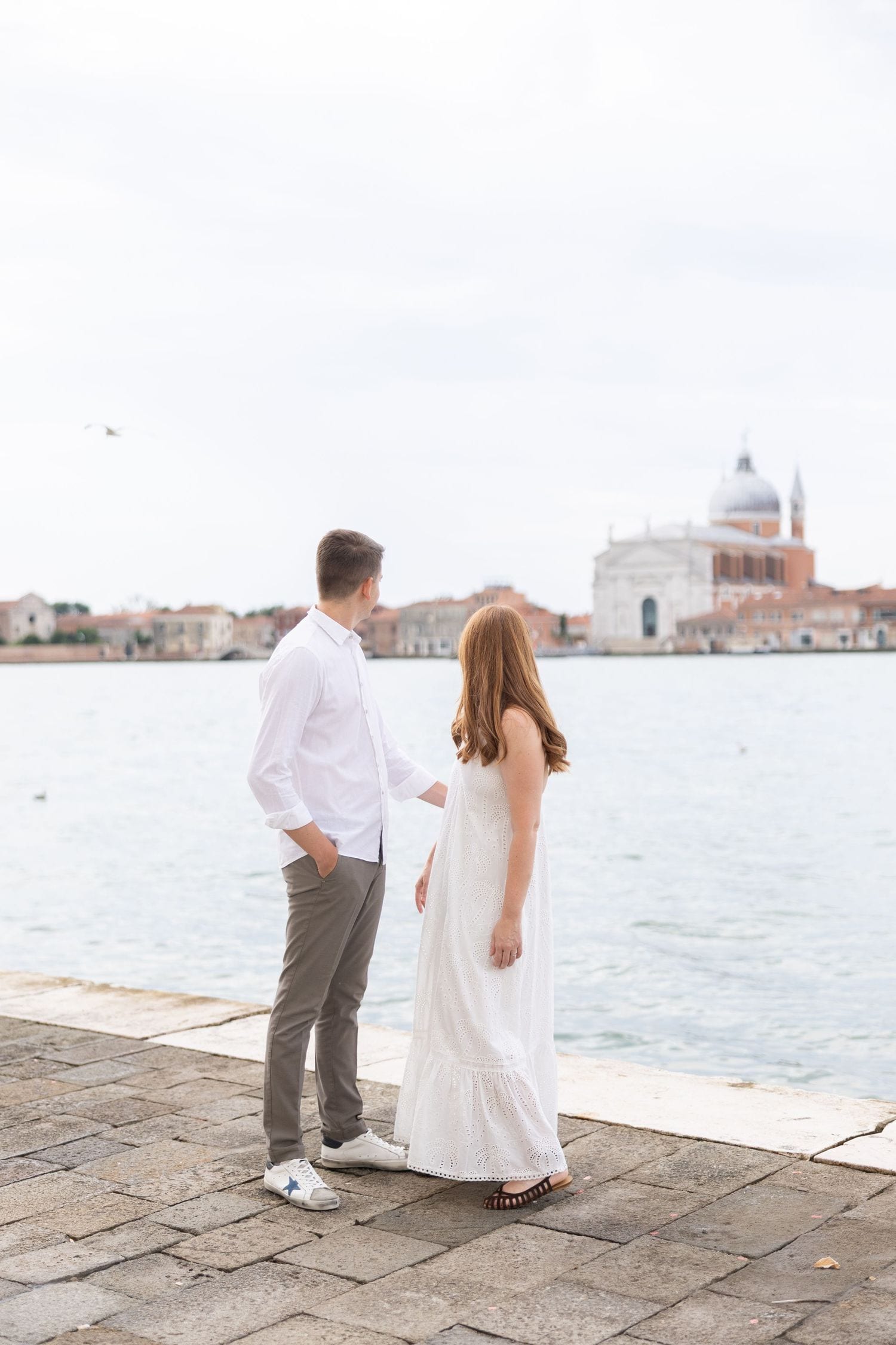 A couple walking along the waterfront promenade with Venice's skyline in the distance.