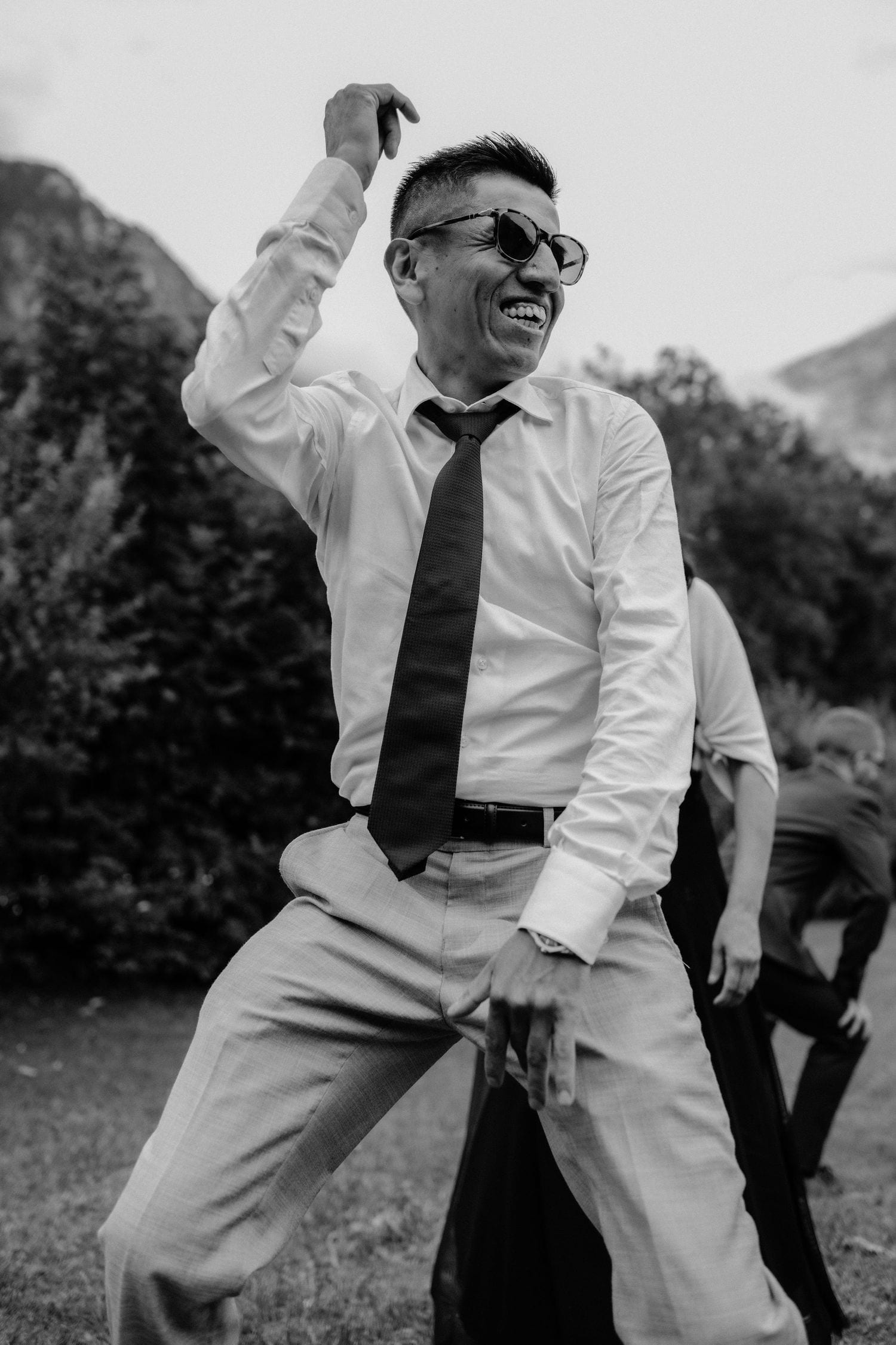Black and white photo of a man in white shirt and tie dancing joyfully outdoors at a celebration.
