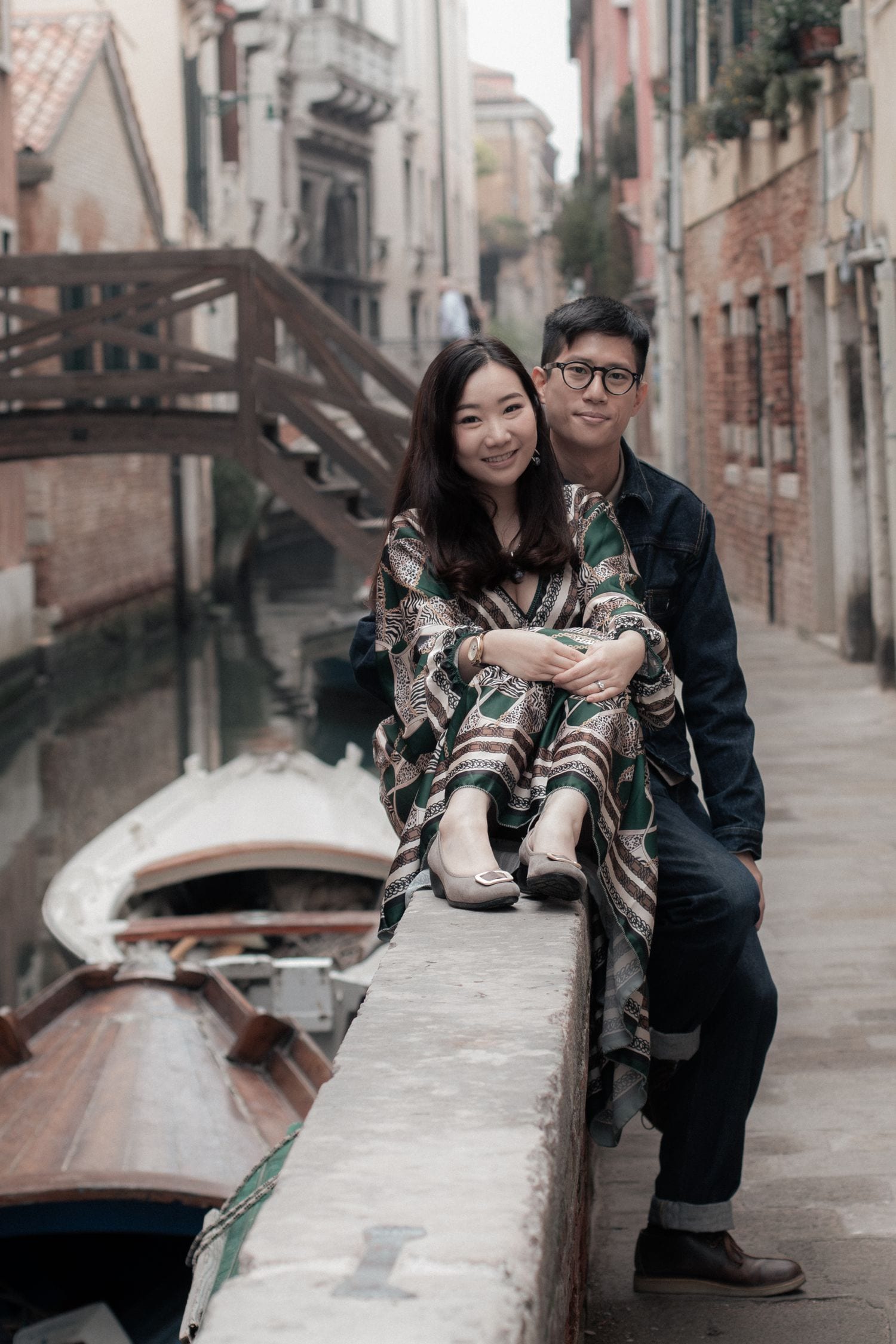 Couple seated beside a quiet canal with boats and historic Venice architecture behind them.