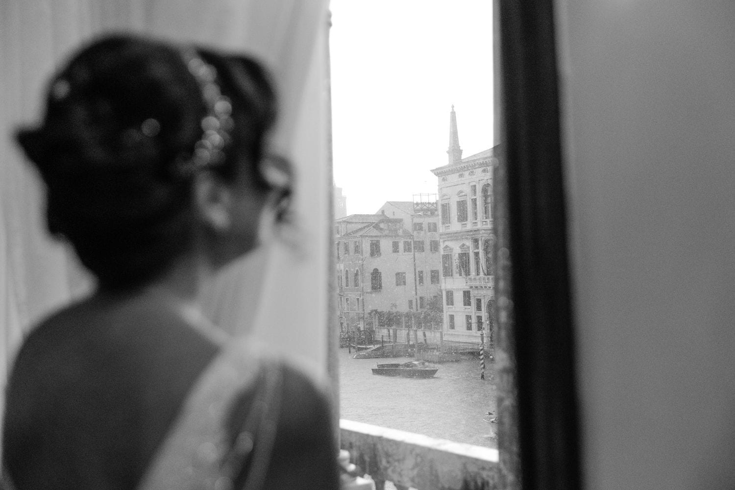 Black and white view through a Venetian window showing a woman silhouetted against the Grand Canal and historic buildings.