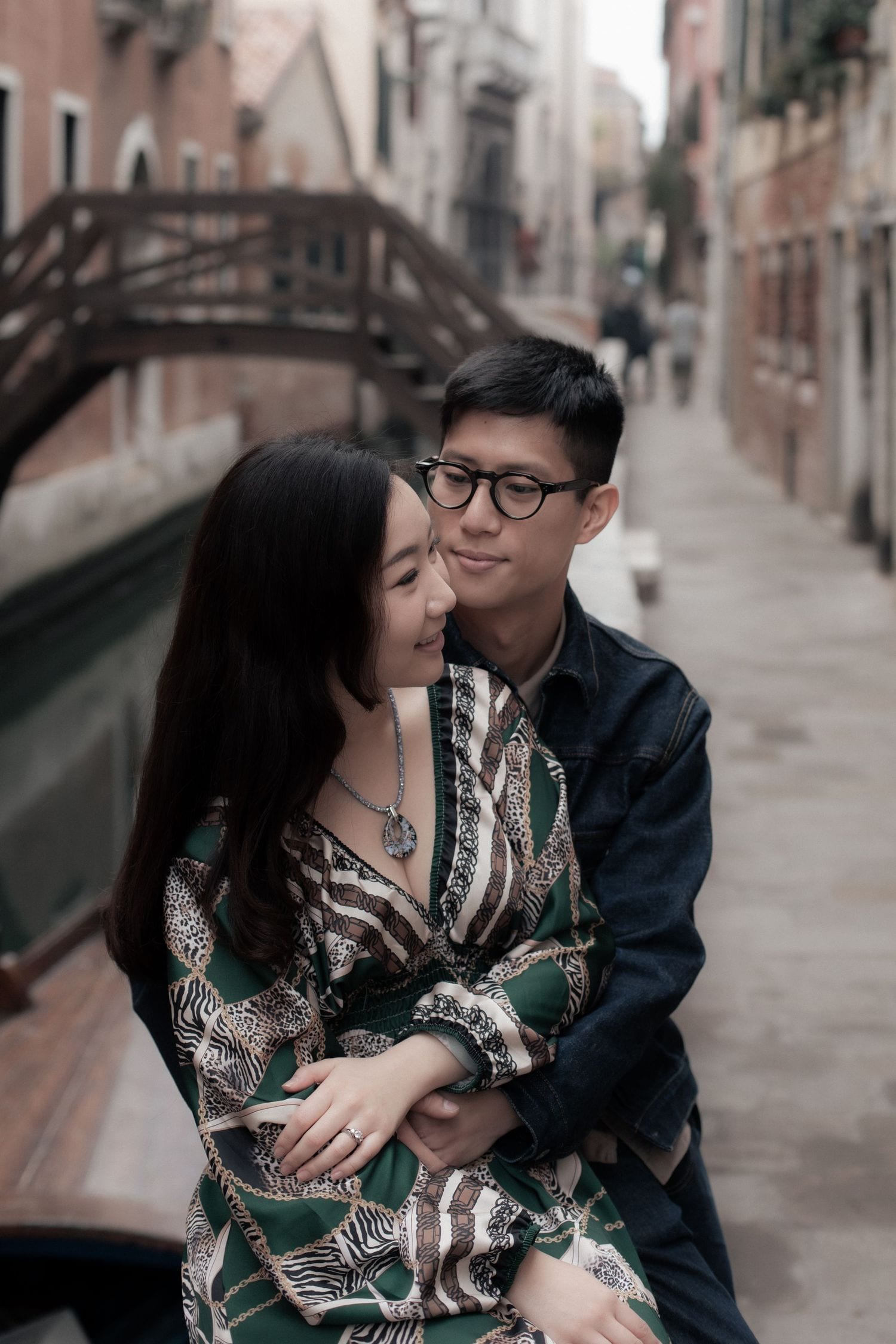 Couple embracing romantically along a Venice canal with historic bridge and buildings in the background.