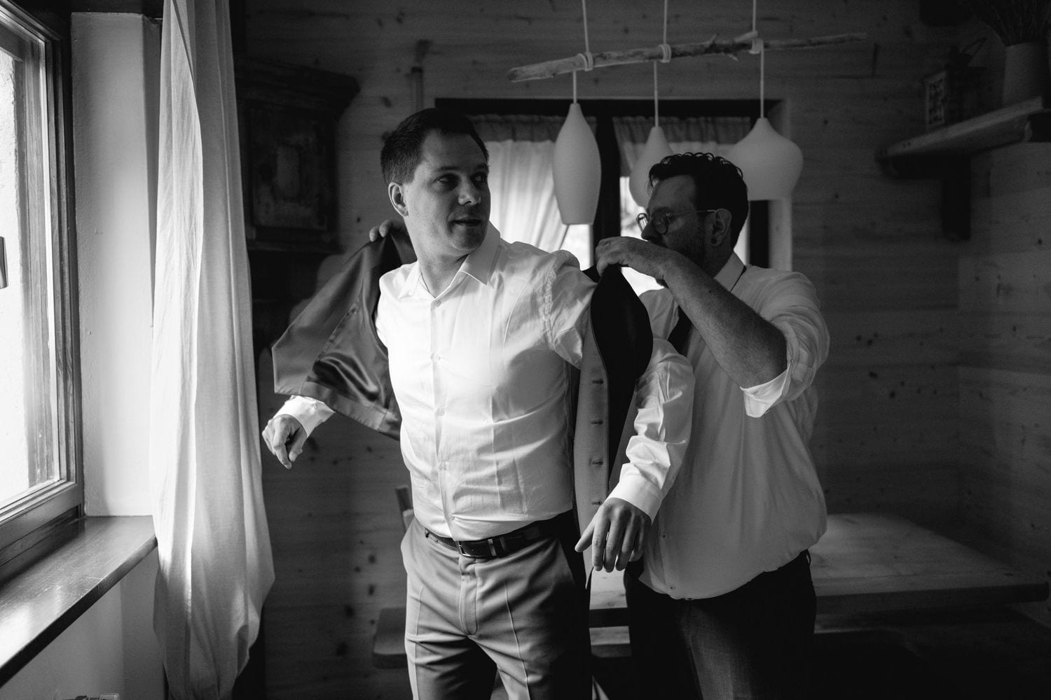 Two men in white dress shirts help each other get dressed in a rustic wooden room in this black and white photo.