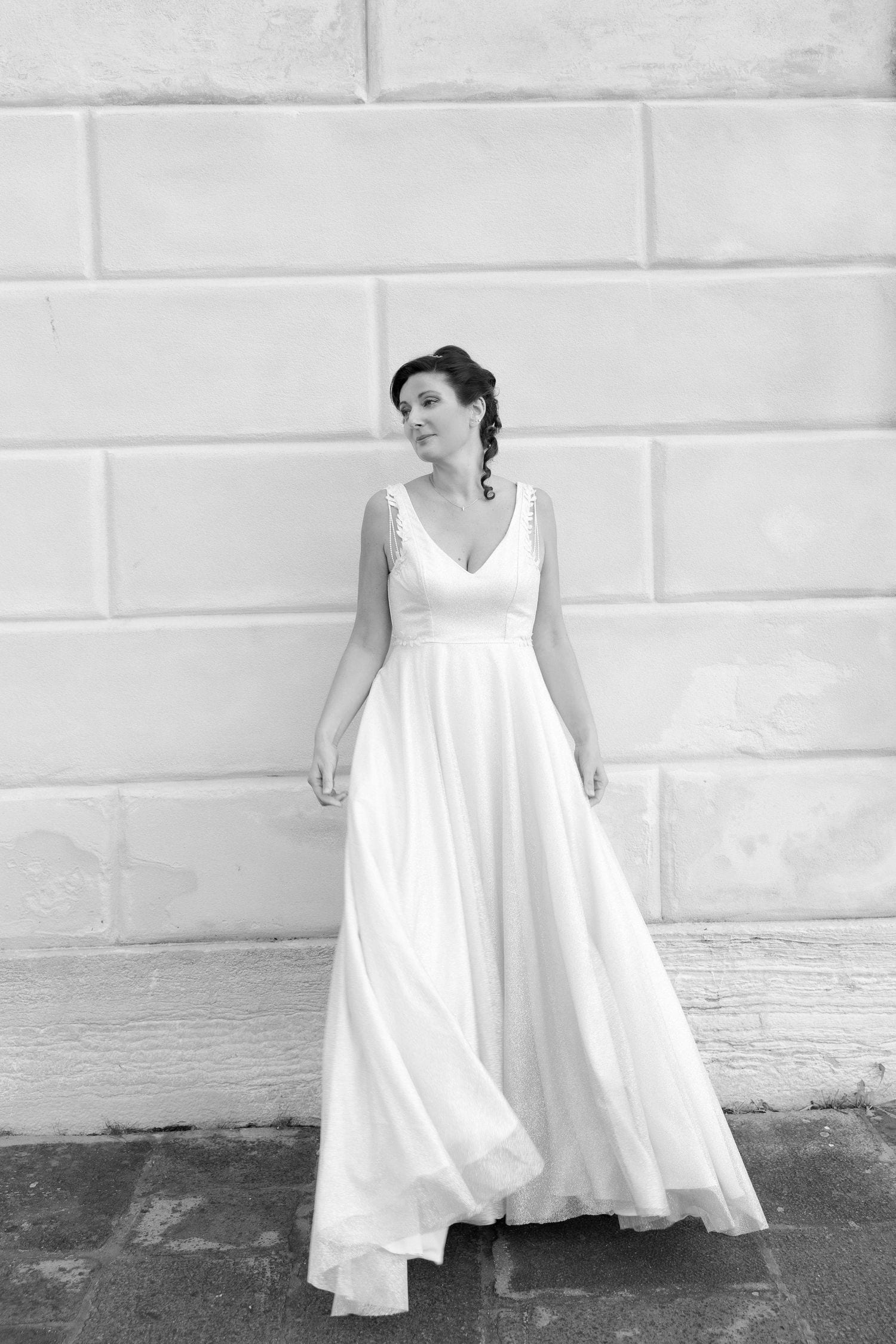 A graceful figure in a flowing white wedding gown poses against a light brick wall in black and white photography.