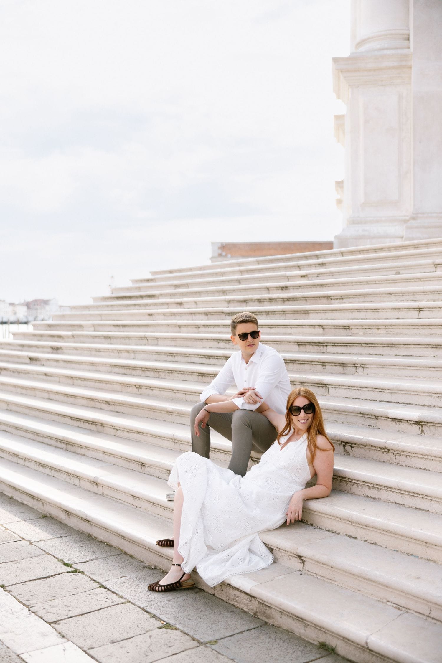A couple in white attire relaxes on grand marble steps of a historic European building on a bright day.