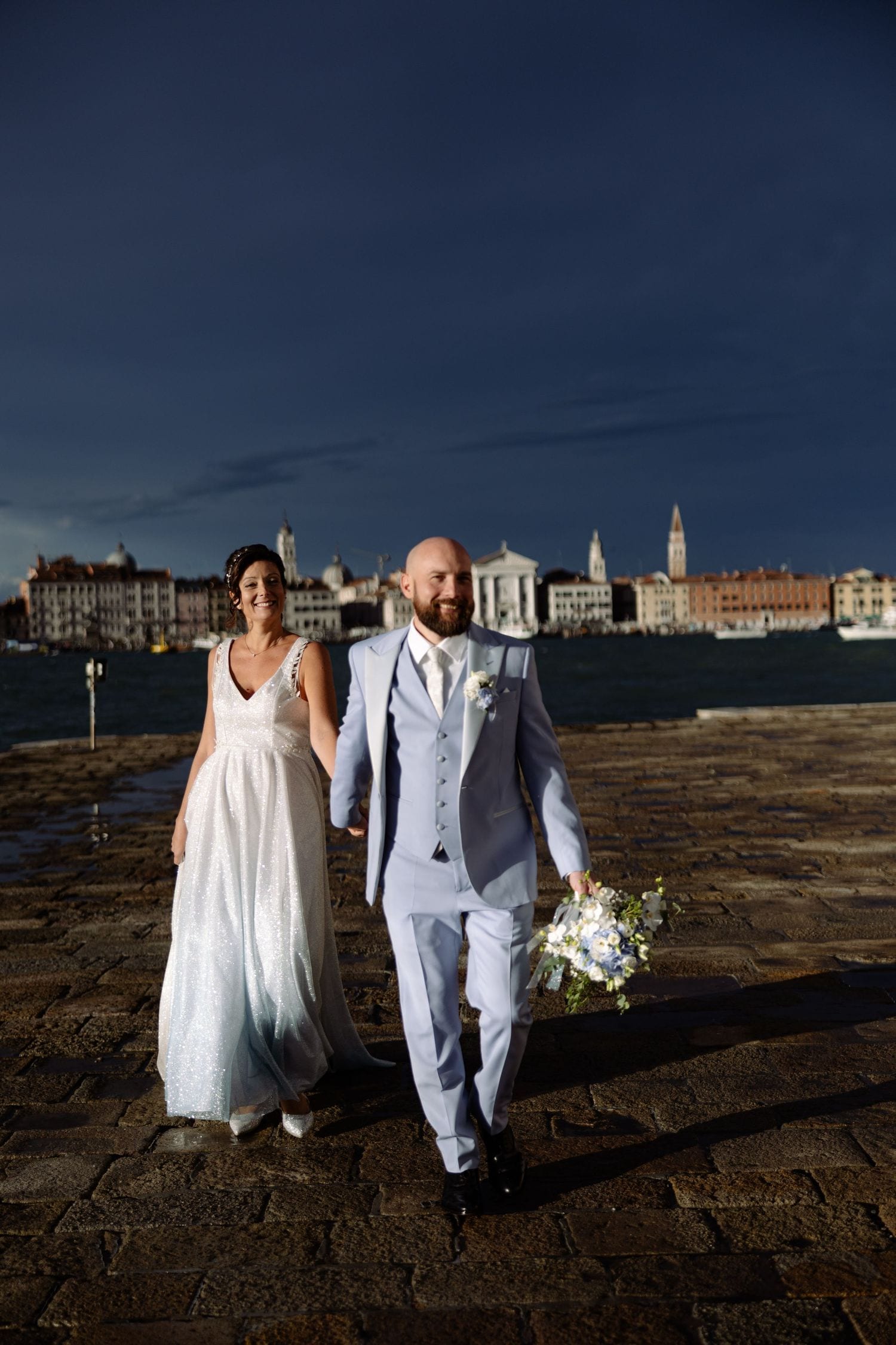 A bride and groom walking along Venice's waterfront with historic buildings in the background.