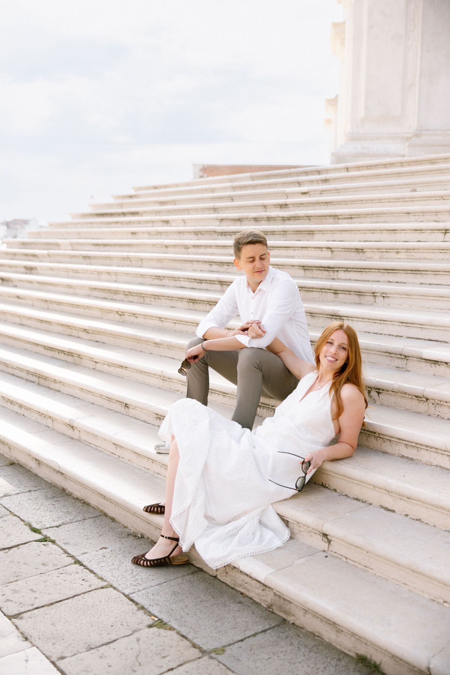 A couple in white outfits relaxes on wide marble steps of a Venetian building.