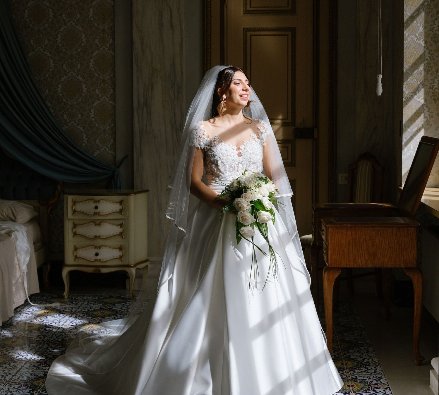 Bride in white ball gown with veil holds bouquet, standing in sunlit elegant room with ornate tiled floor.