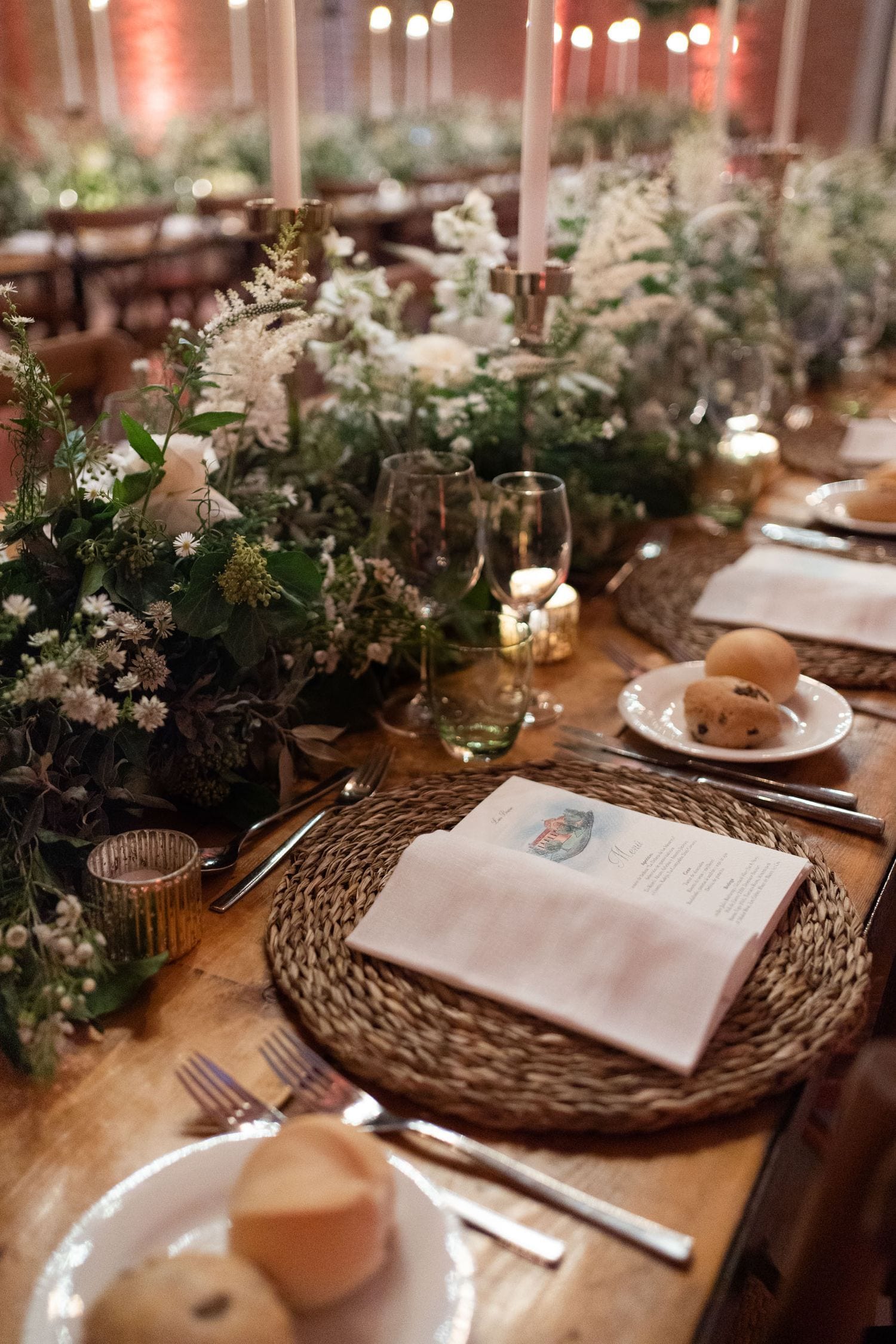 Elegant wedding reception table with woven placemats, white floral centerpieces, candles, wine glasses, and menu cards on wooden table.