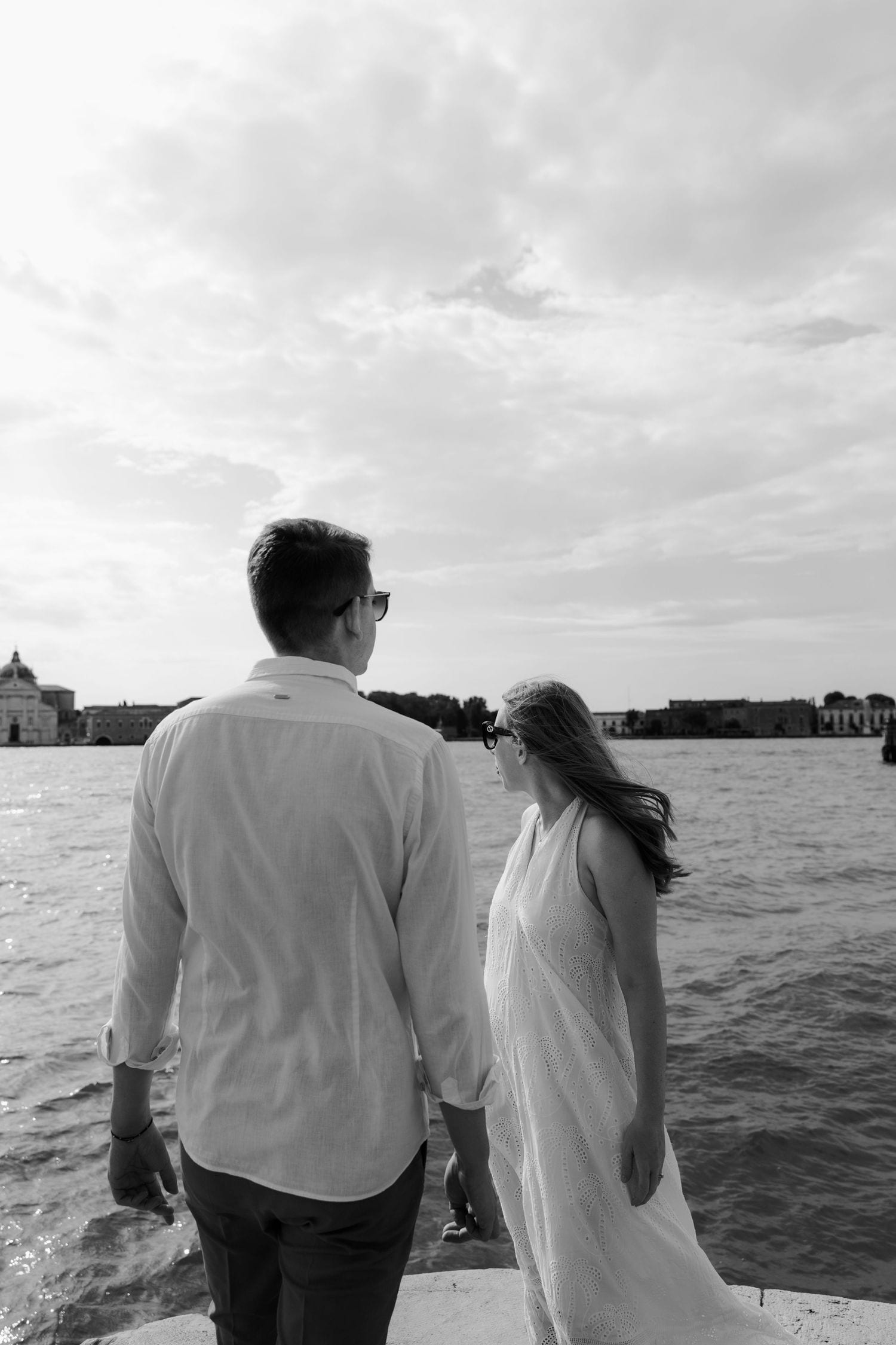 A couple looks out over Venice's lagoon on a cloudy day with church domes visible in the distance.