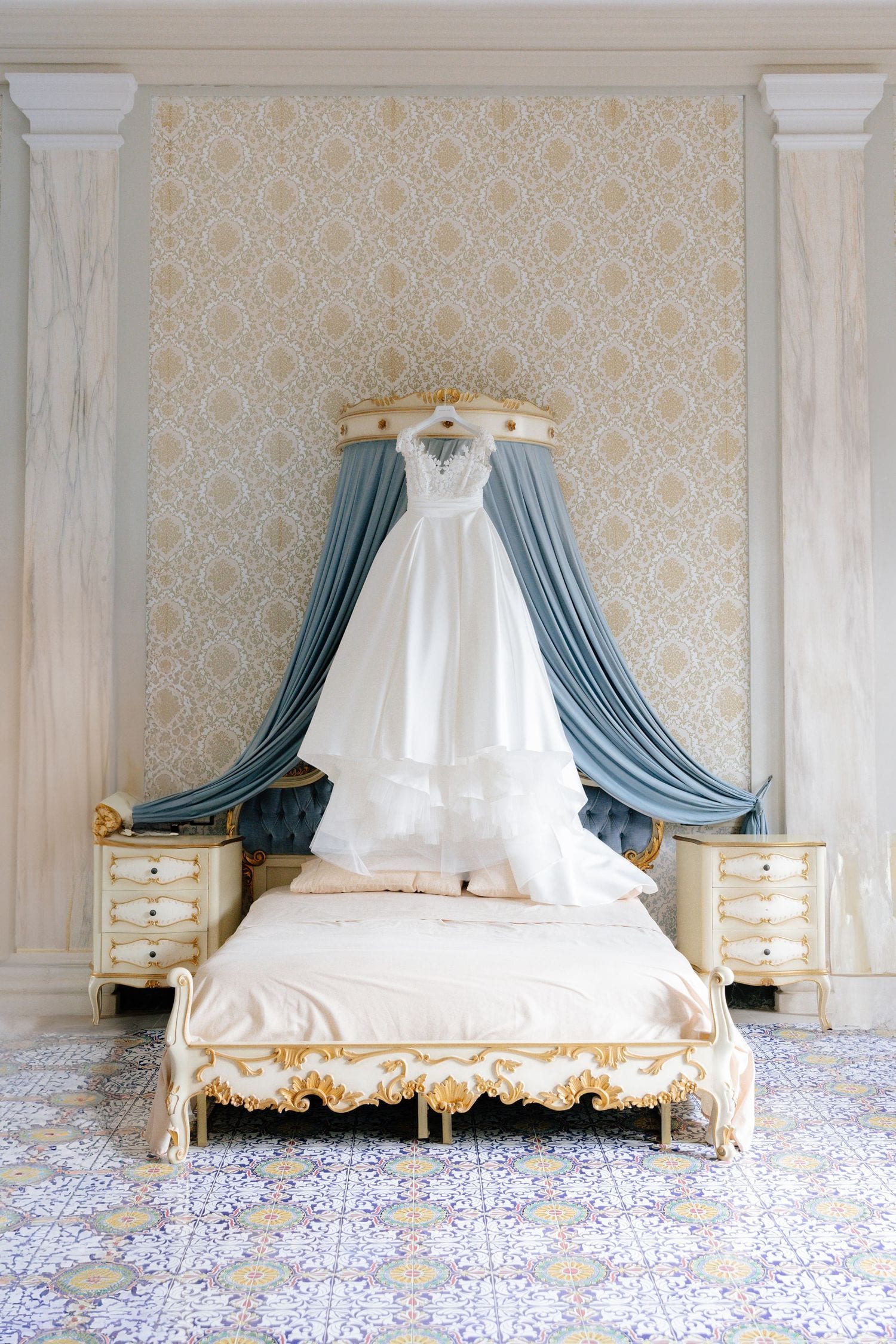 Elegant white wedding dress hanging on a ornate canopy bed with blue drapes in a luxurious vintage bedroom.