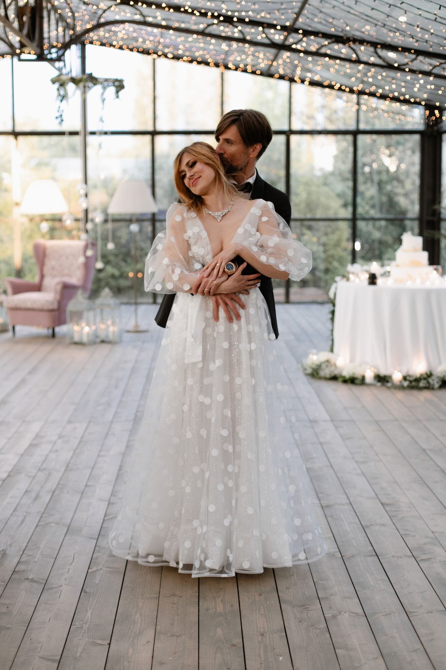 A couple shares an intimate moment during their wedding celebration in a rustic greenhouse venue with string lights overhead.