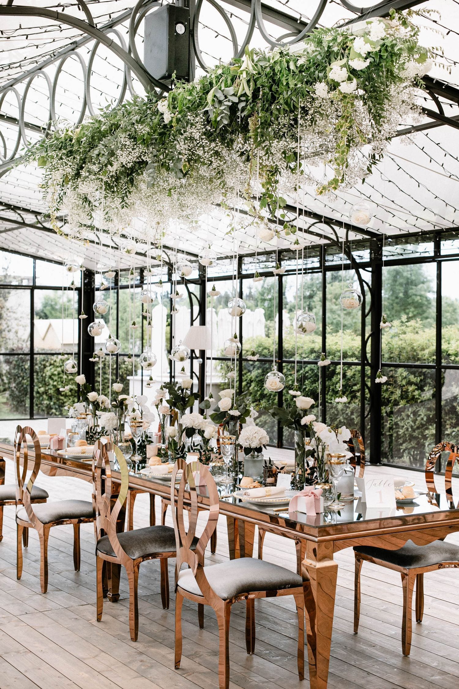 Elegant greenhouse wedding reception with wooden tables, hanging greenery and natural light streaming through glass ceiling.