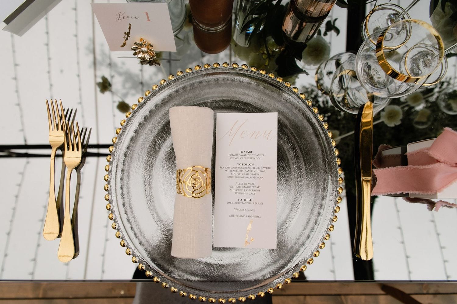 Elegant table setting with gold cutlery and beaded silver charger plate featuring crisp white menu card at wedding reception.