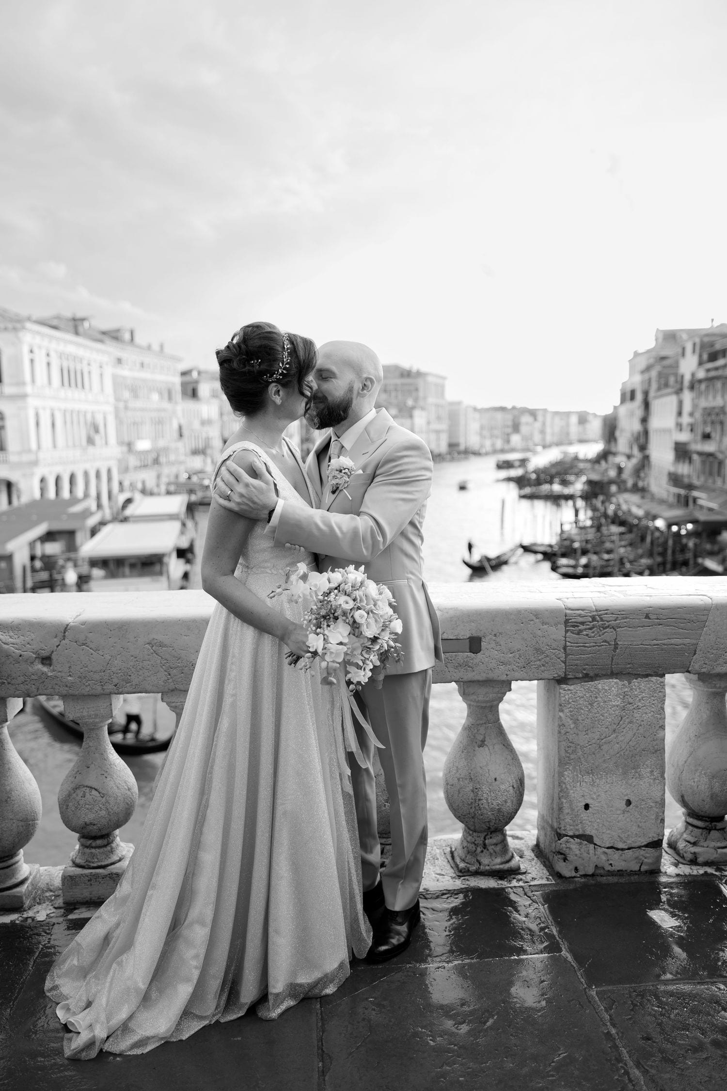 A romantic wedding couple embraces on a stone balcony overlooking Venice's iconic Grand Canal in black and white.