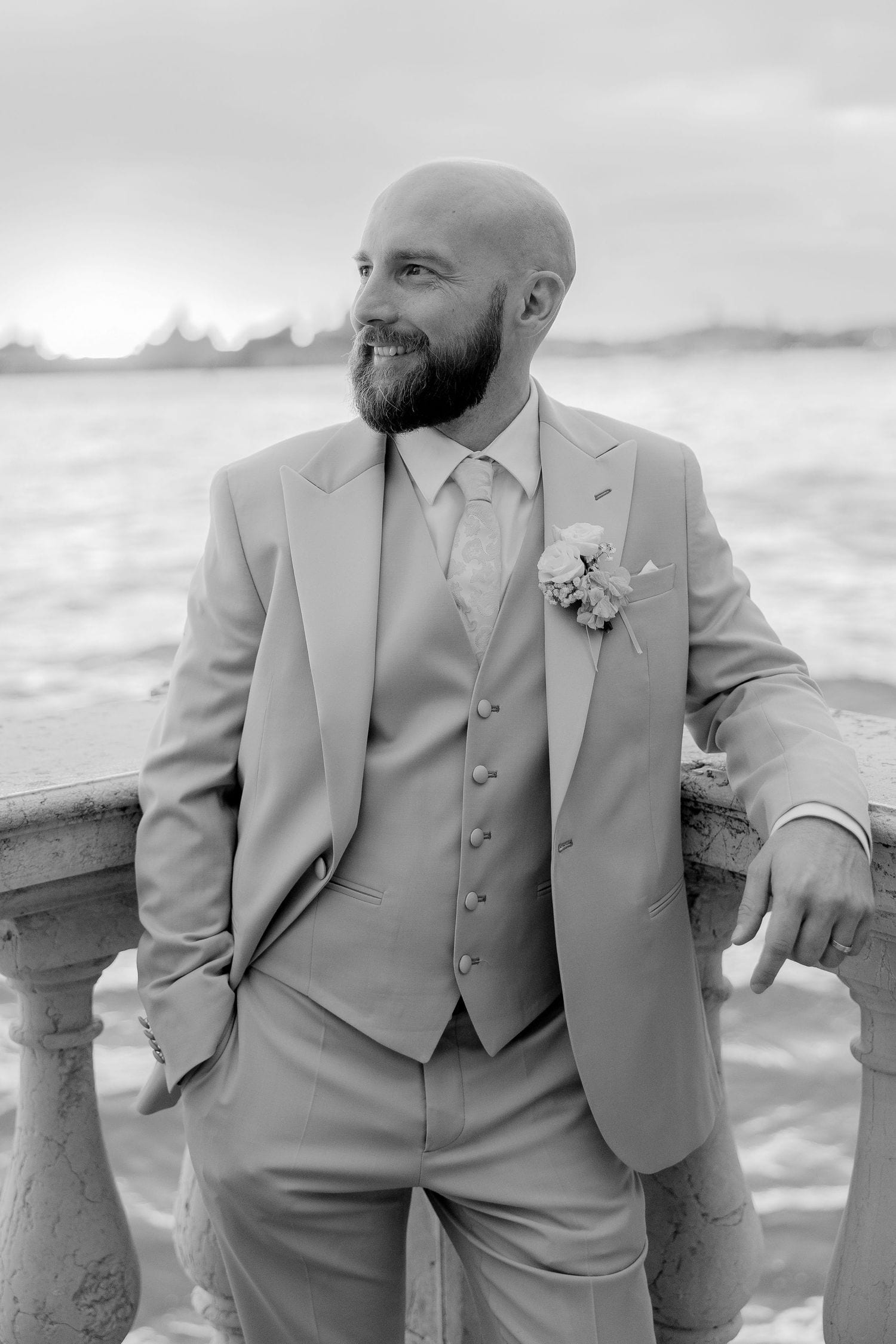 A groom in a light gray three-piece suit poses by a stone railing with an ocean backdrop.