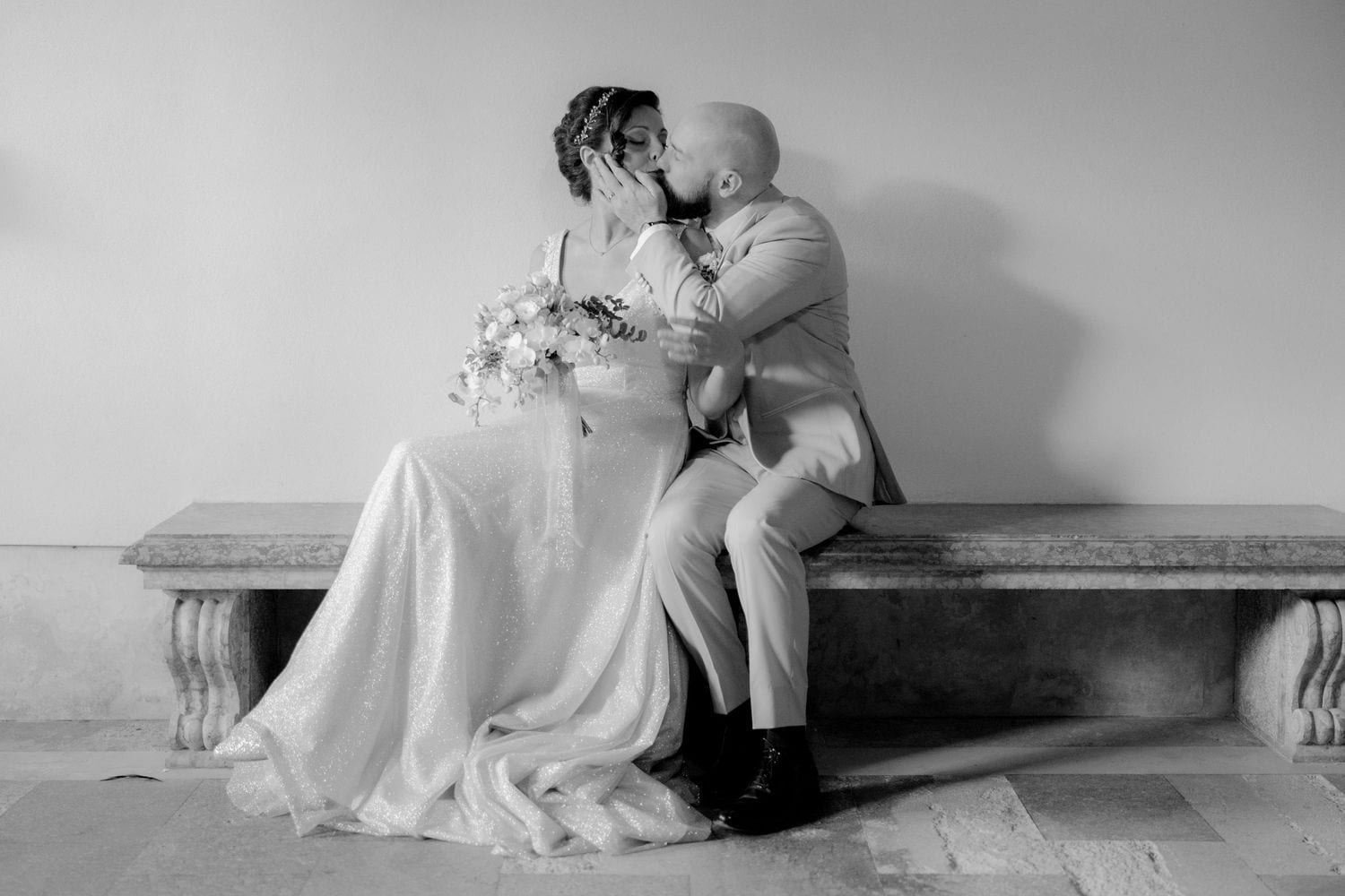 A romantic black and white photo of a couple in wedding attire sharing an intimate moment on a stone bench.