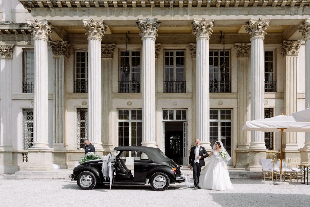 Bride and groom pose beside a vintage black car in front of a grand neoclassical building with tall columns.