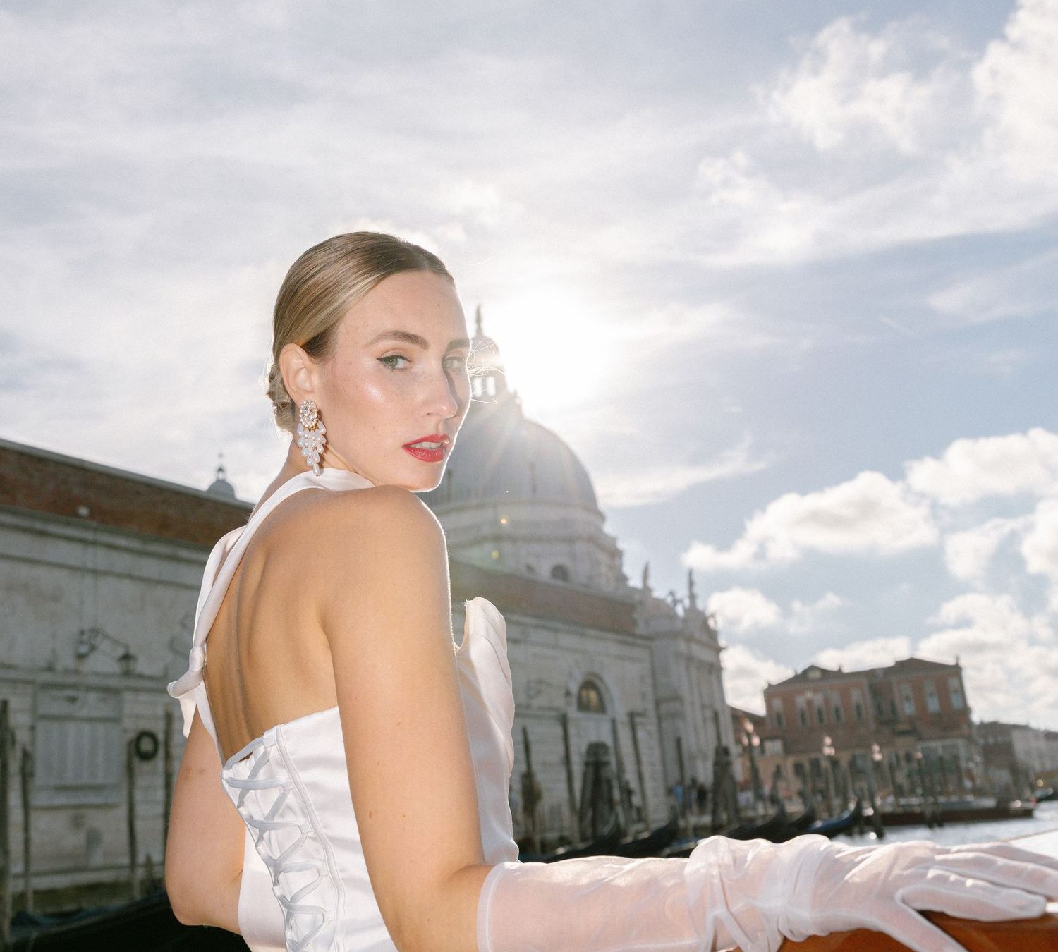 A bride in a white wedding dress poses on a boat in Venice with historic architecture and cloudy skies in the background.