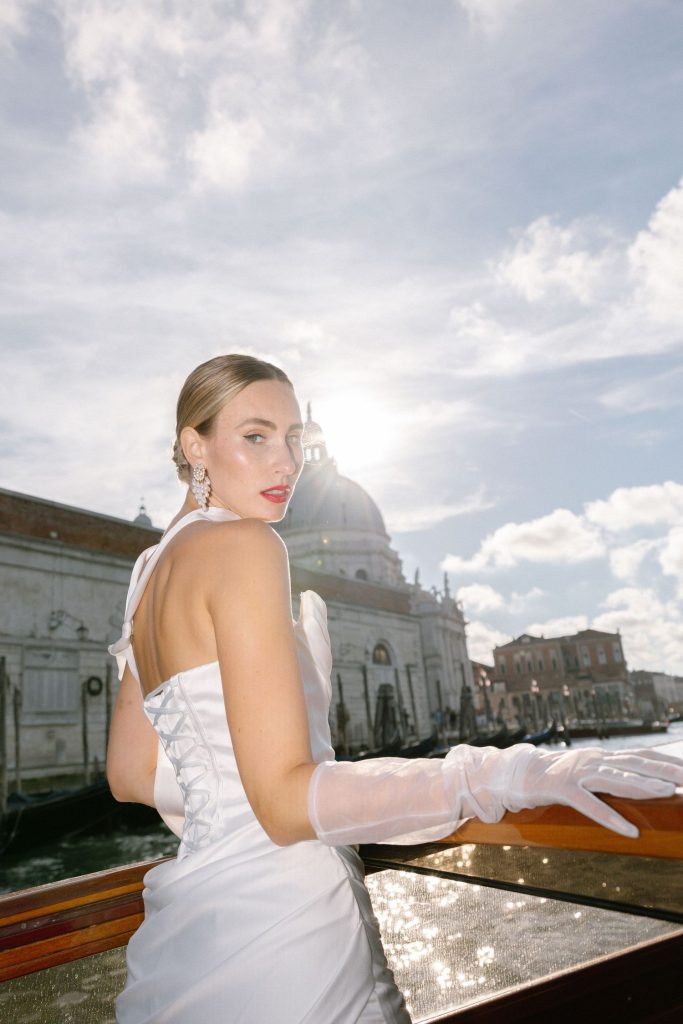 A bride in a white wedding dress poses on a boat in Venice with historic architecture and cloudy skies in the background.