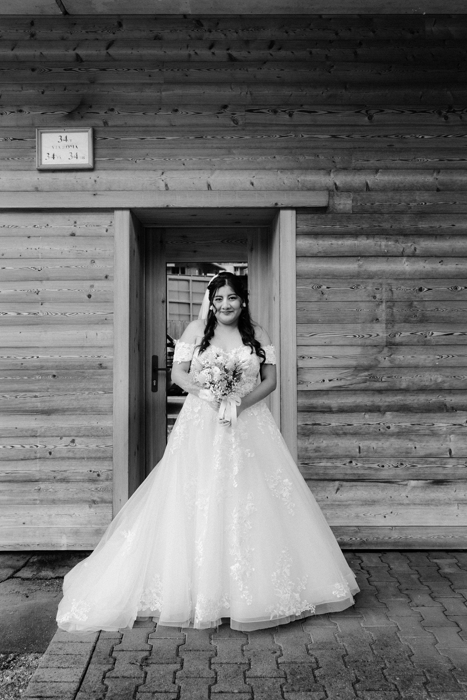 Black and white photo of a bride in a ball gown holding a bouquet, standing in the doorway of a wooden cabin.
