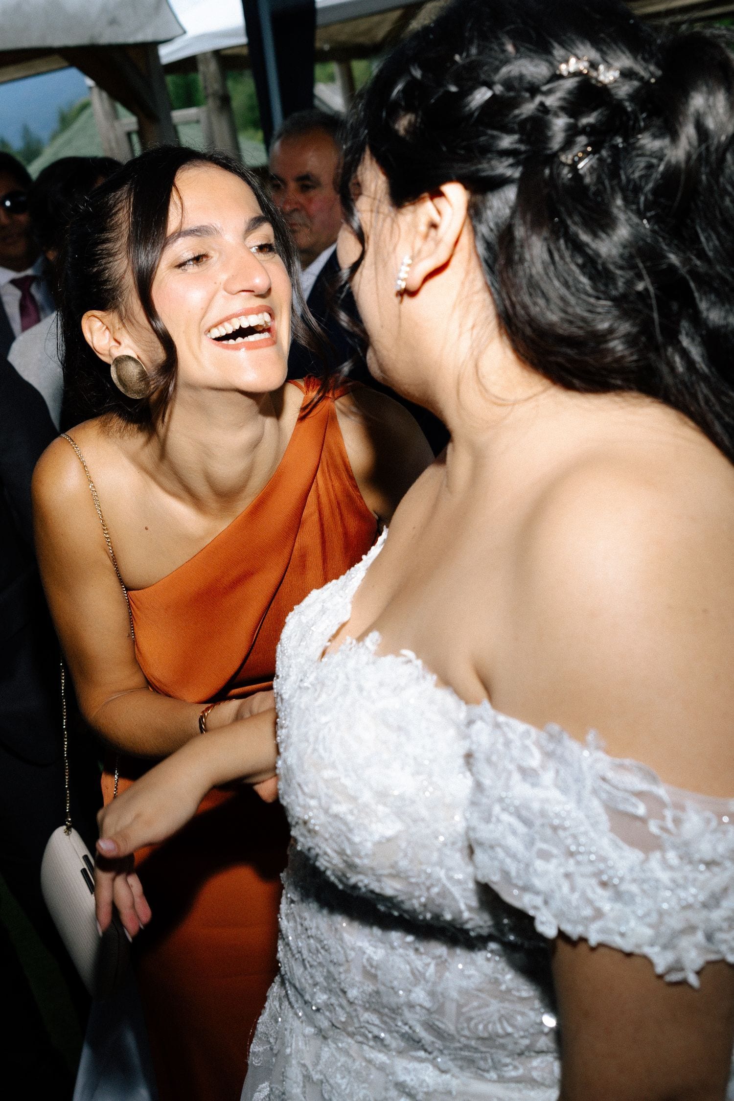 Bride in white lace gown laughing joyfully with bridesmaid in orange dress at wedding reception.