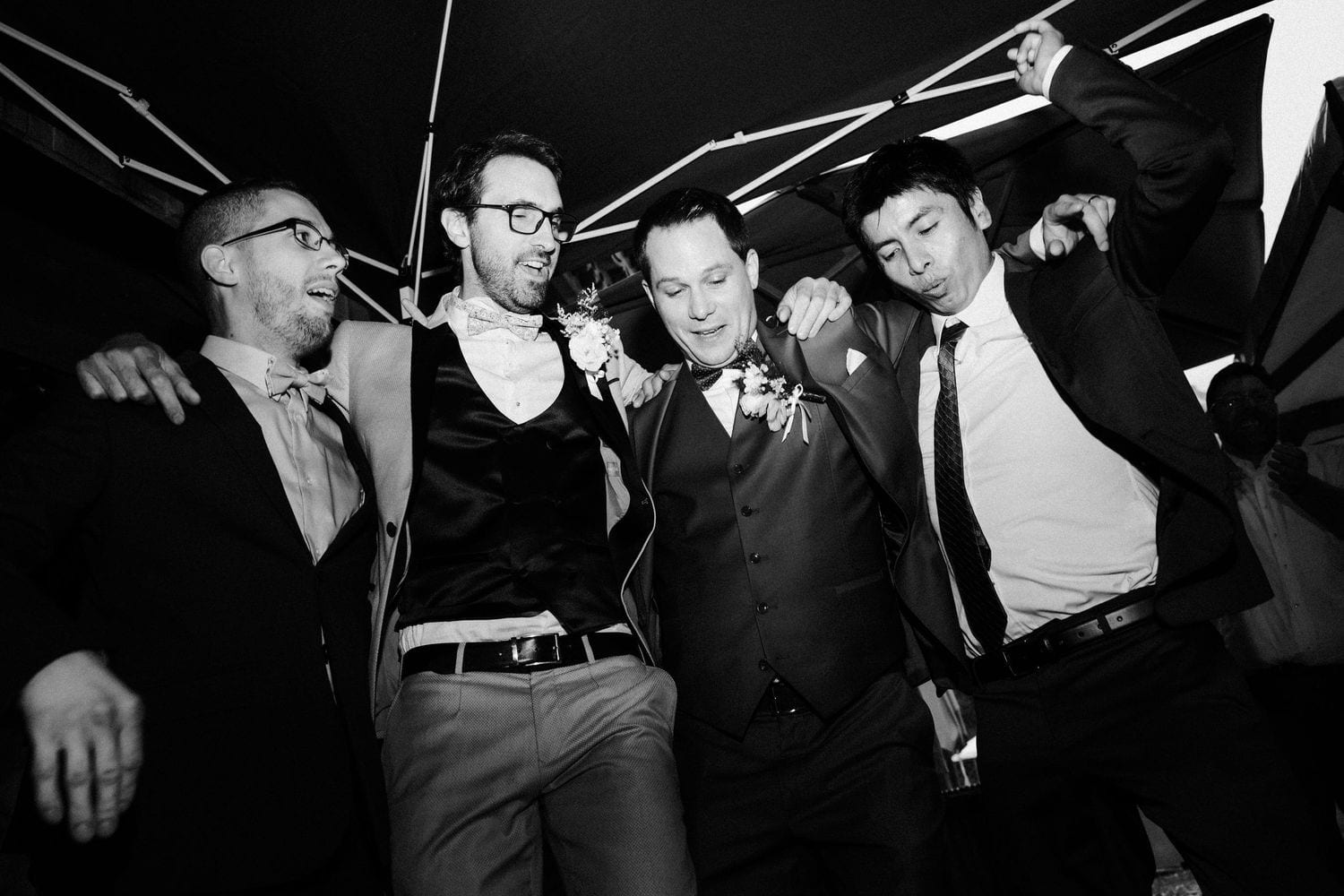 Black and white photo of four men in formal attire celebrating together at a wedding reception.
