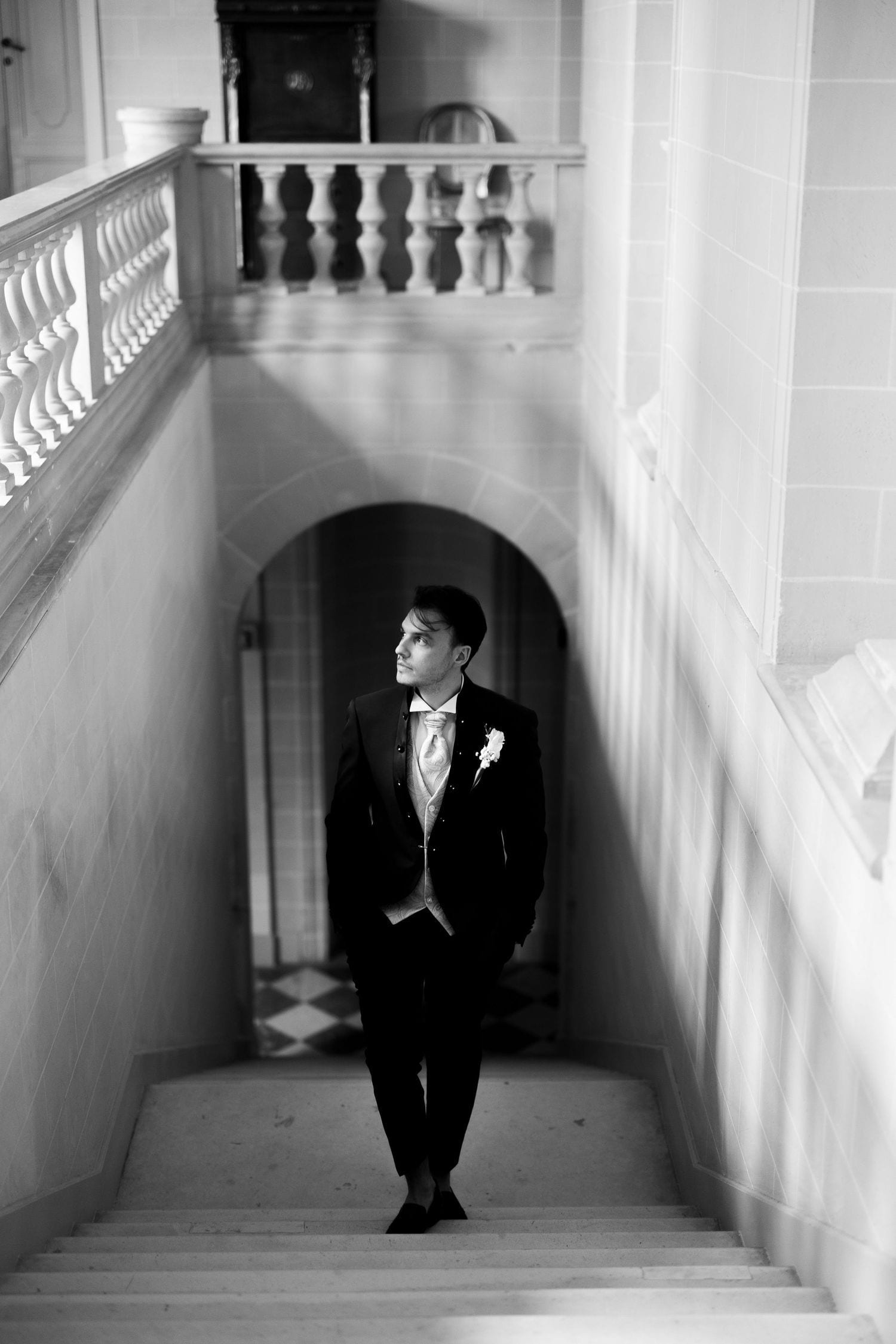 Groom in black tuxedo stands on grand staircase in elegant venue, black and white wedding portrait.