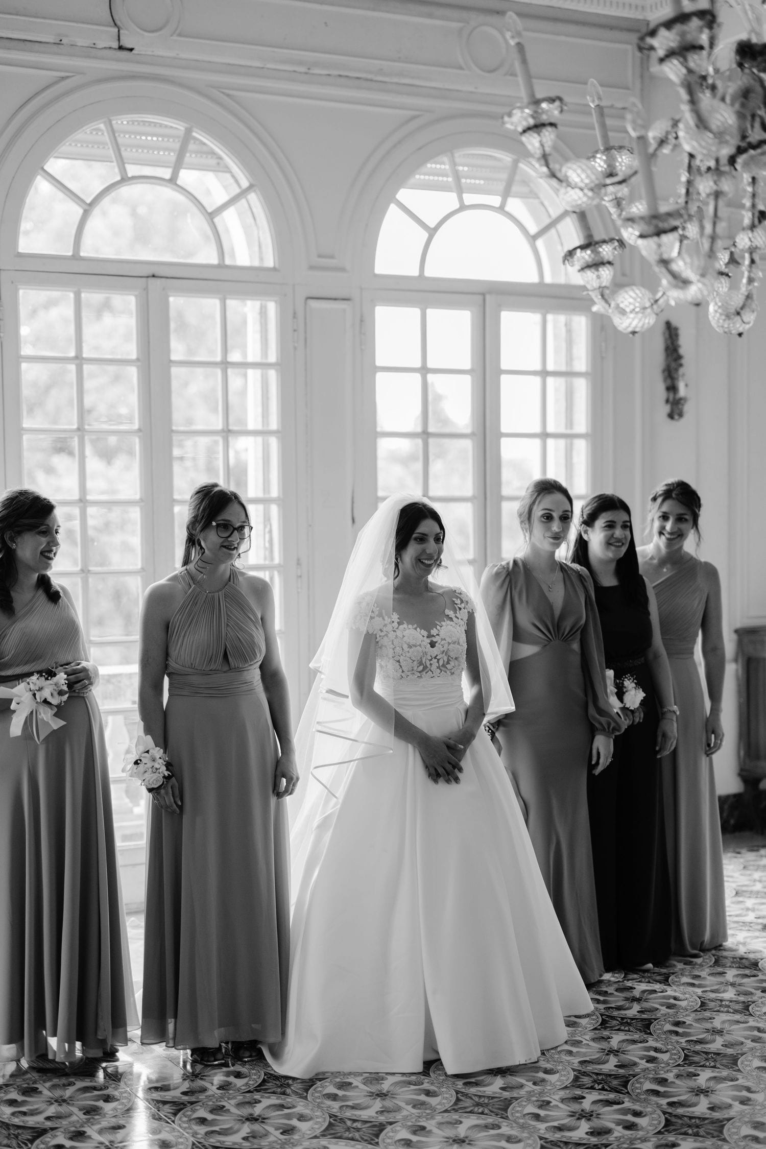 Bride in white gown and veil poses with bridesmaids in matching dresses inside elegant venue, black and white photo.
