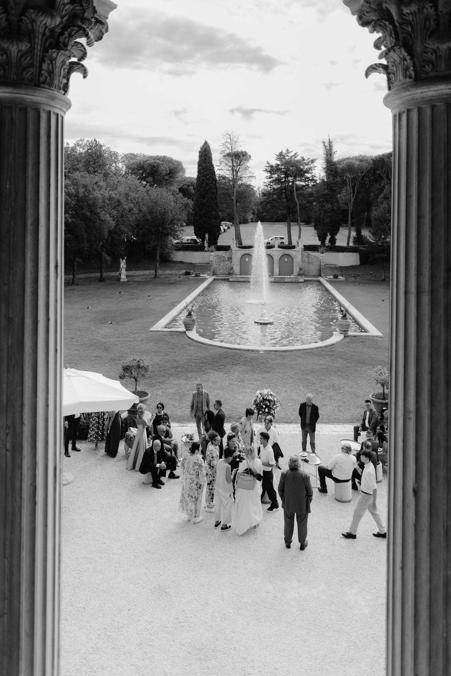 Black and white photo of elegant wedding guests gathered near a fountain in a grand villa garden framed by columns.