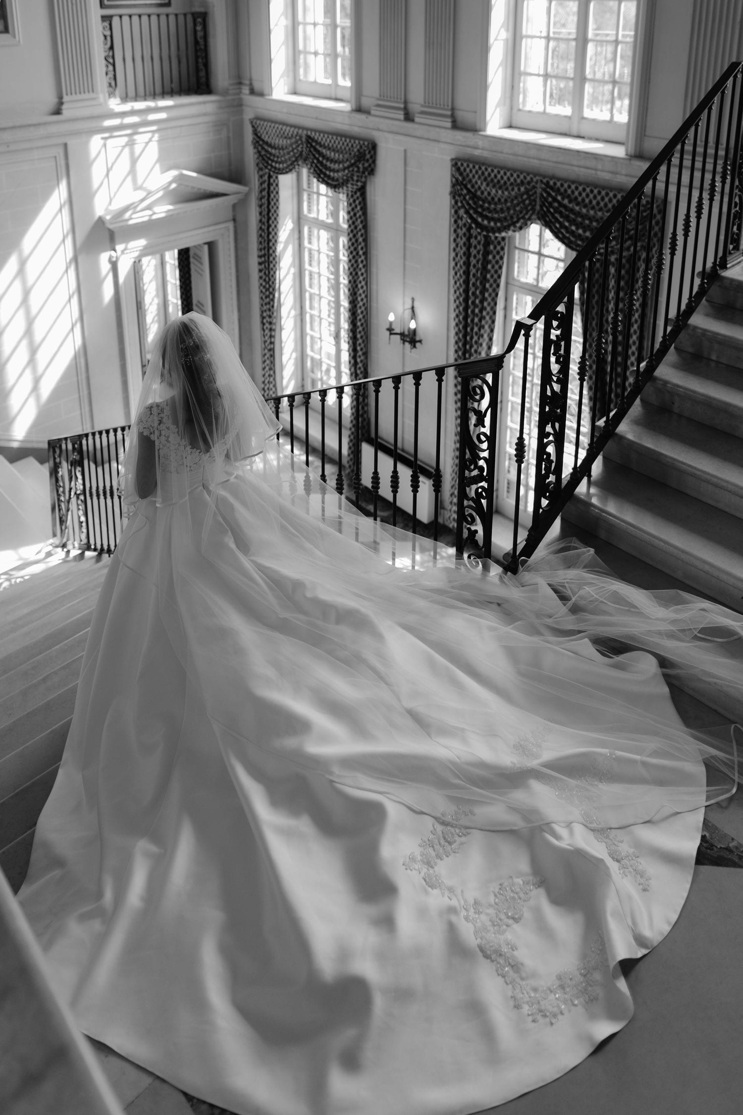 Black and white photo of a bride in a flowing gown and veil descending an elegant staircase in a grand mansion.