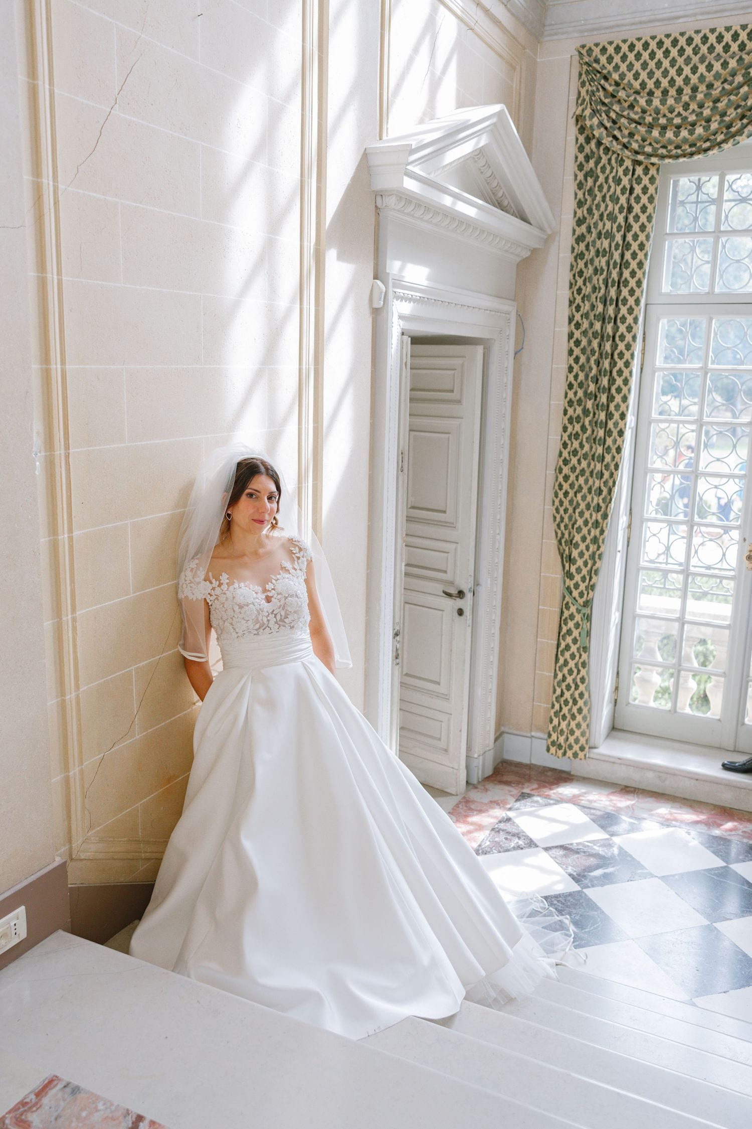 Bride in a white lace ballgown and veil poses elegantly in a sunlit classical interior with marble floors.