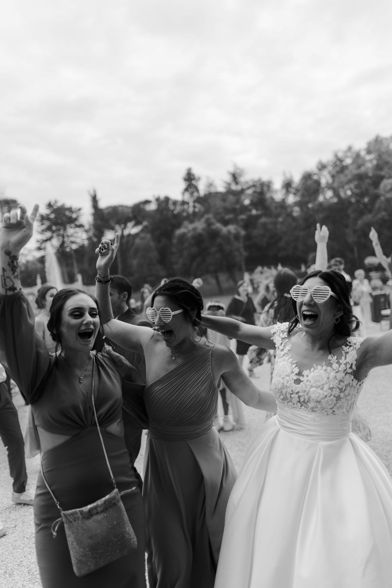 Black and white photo of a bride in white gown and sunglasses celebrating joyfully with bridesmaids at outdoor wedding.