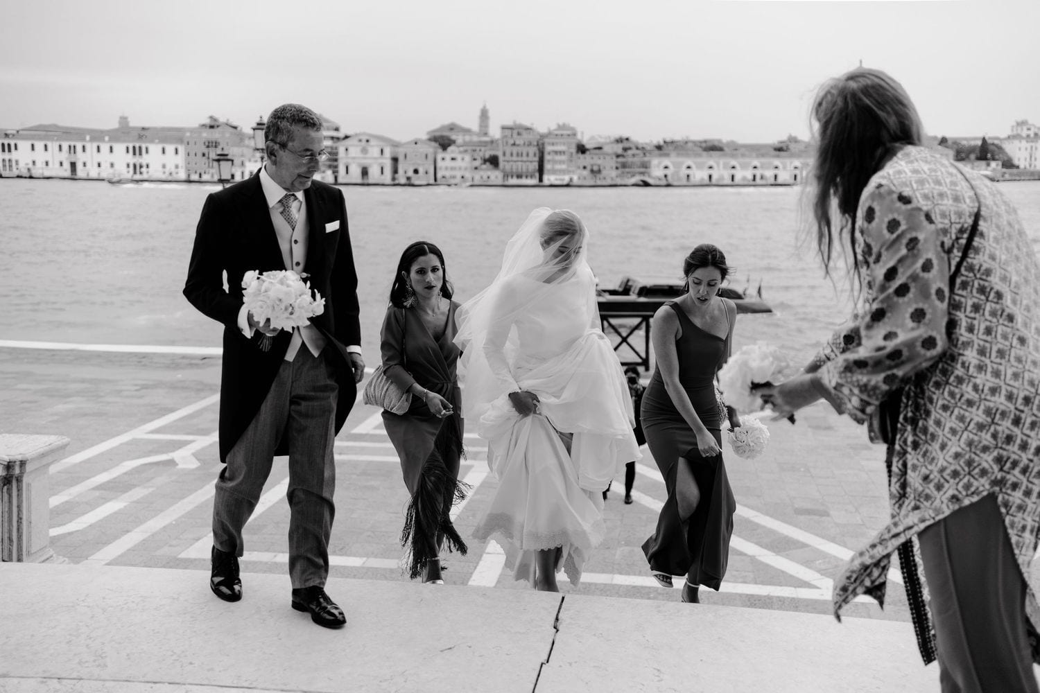 Black and white photo of a wedding entrance at the Church walking near waterfront in Venice, Italy, with bride in veil.