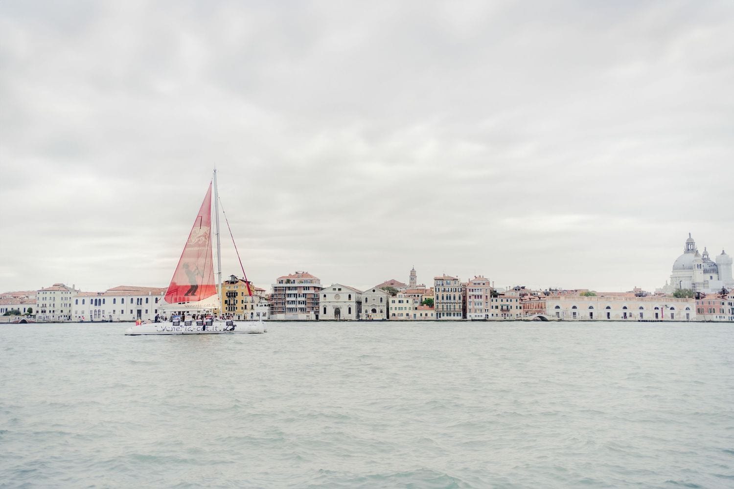Red sailboat gliding past the historic Venice skyline with the Santa Maria della Salute dome under overcast skies.