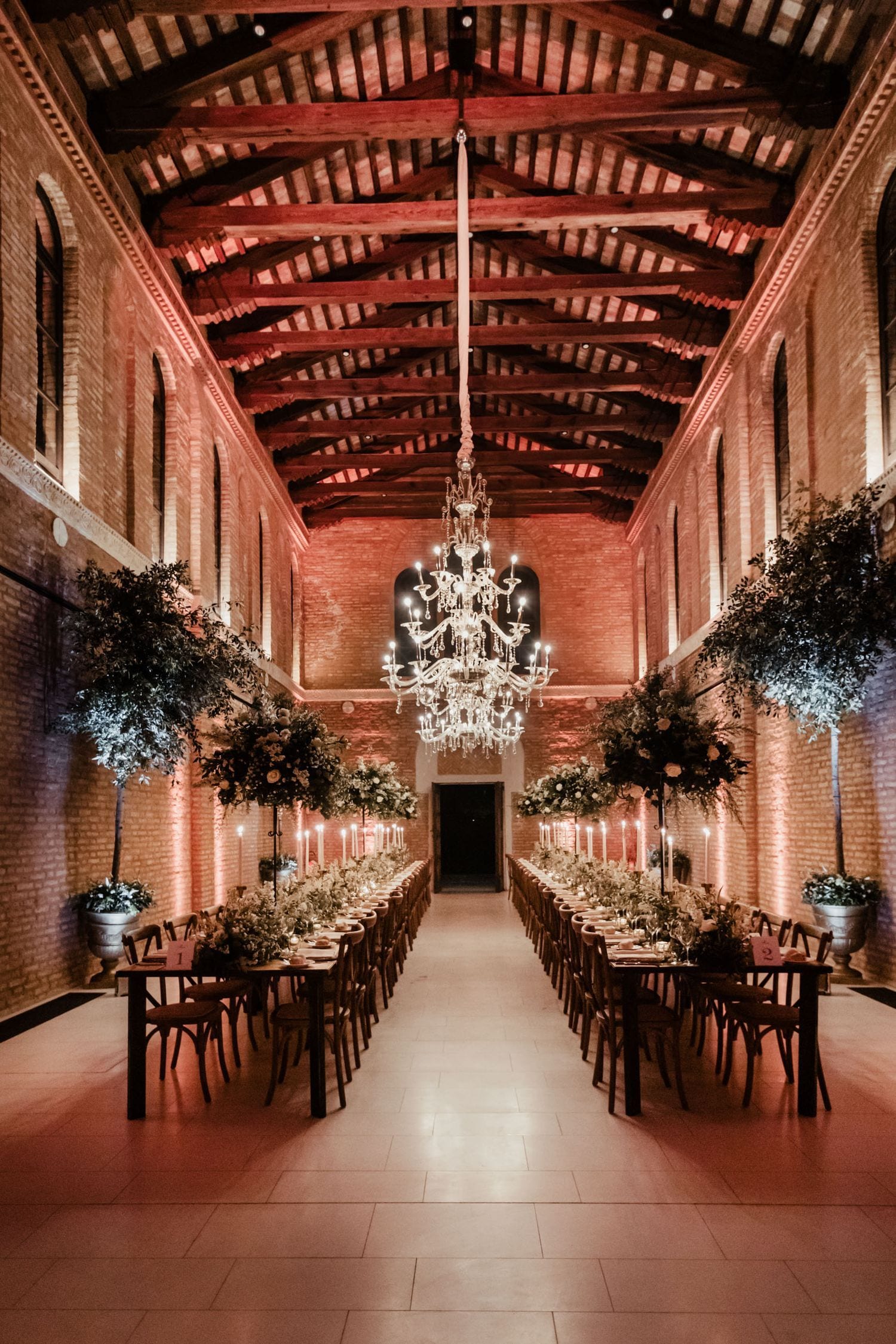 Elegant wedding reception in brick hall with crystal chandelier, long dining tables, floral arrangements, and warm amber lighting.