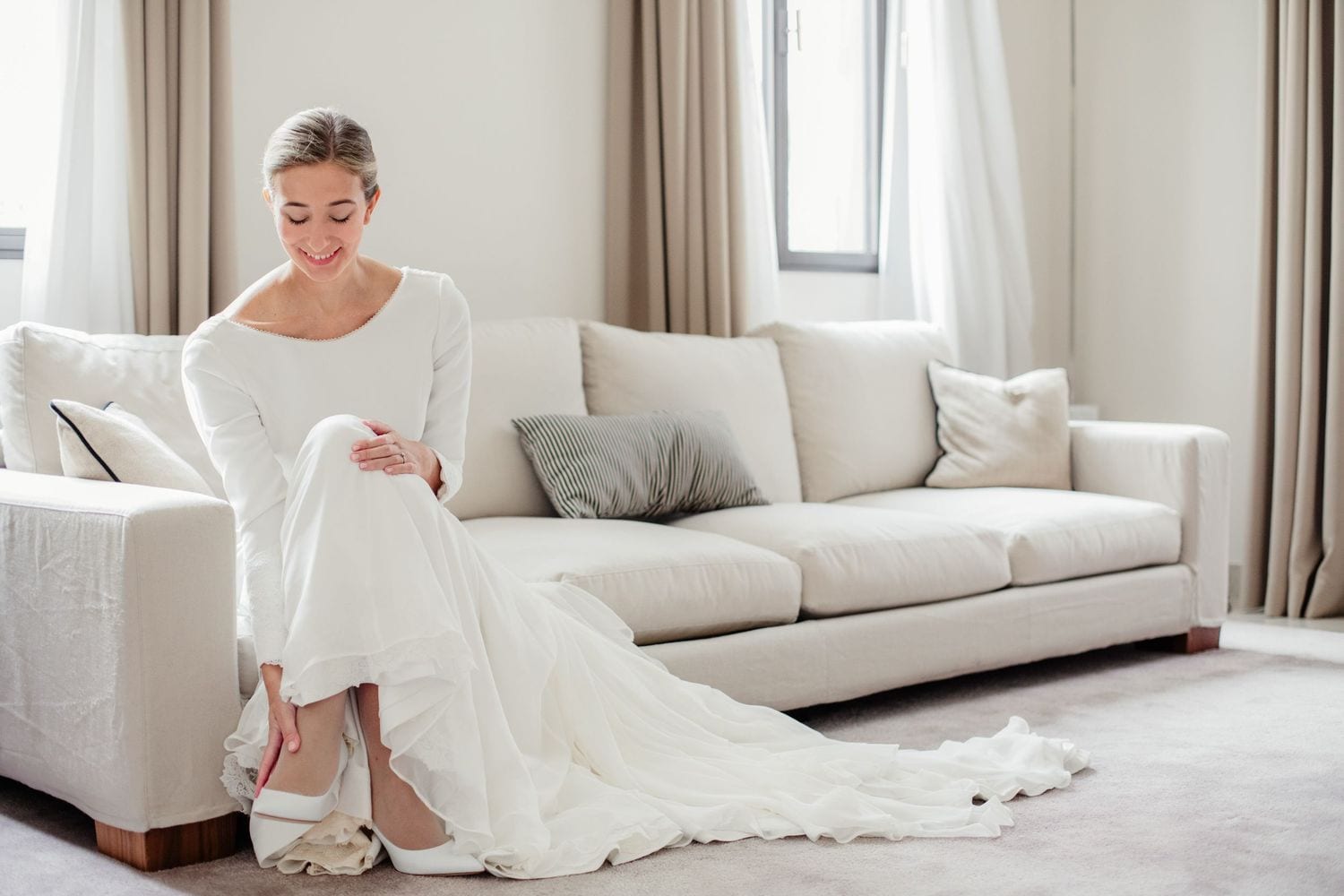 Bride in elegant white wedding dress sitting on cream sofa, adjusting her bridal heels in a bright, modern room.