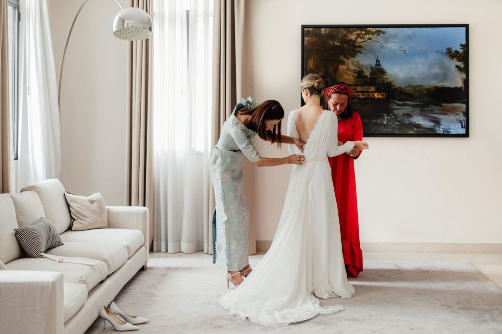 Bride in white gown being helped by two women in elegant room with classic painting backdrop.