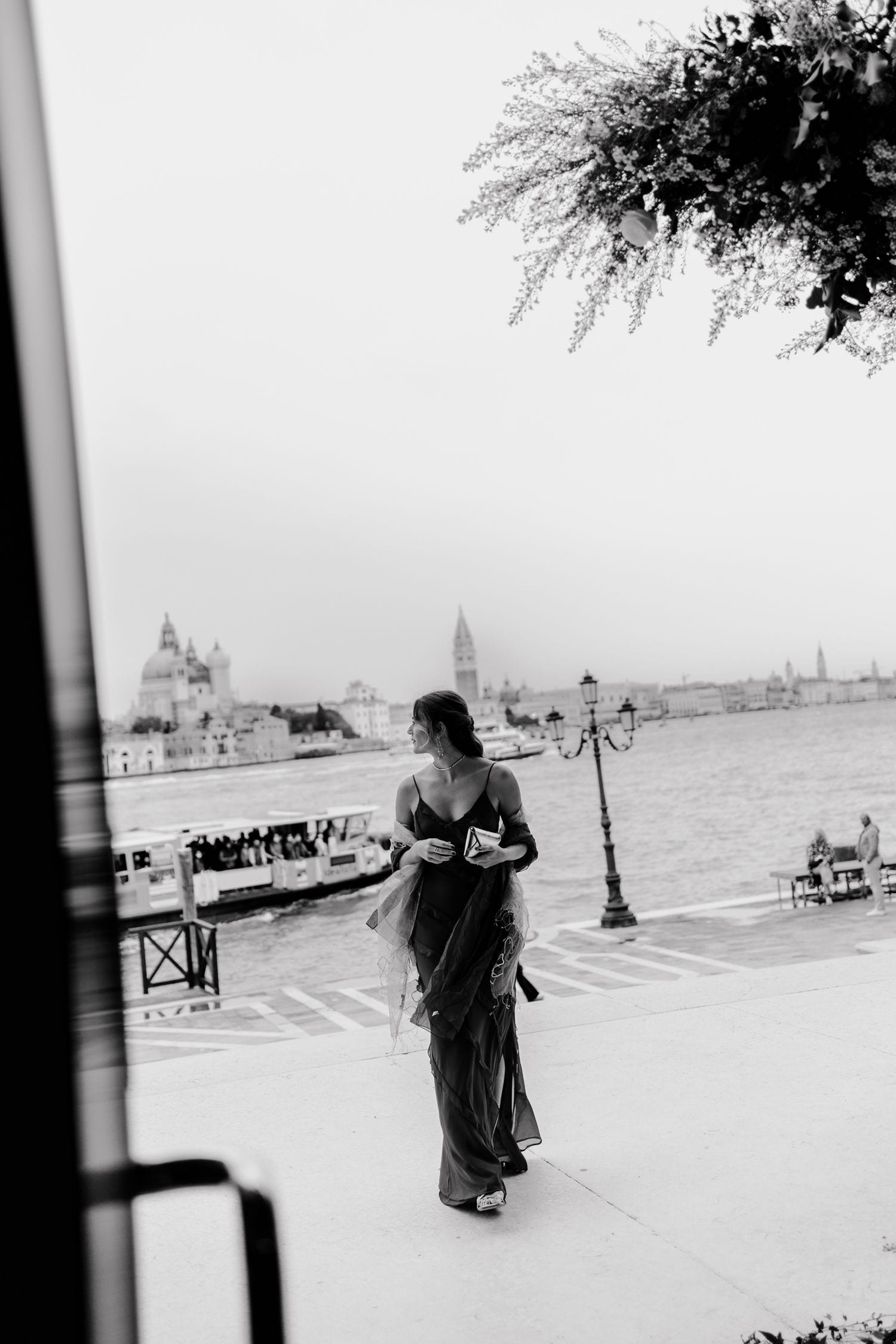 Black and white photo of an elegant woman in a flowing gown walking along a Venice waterfront promenade.